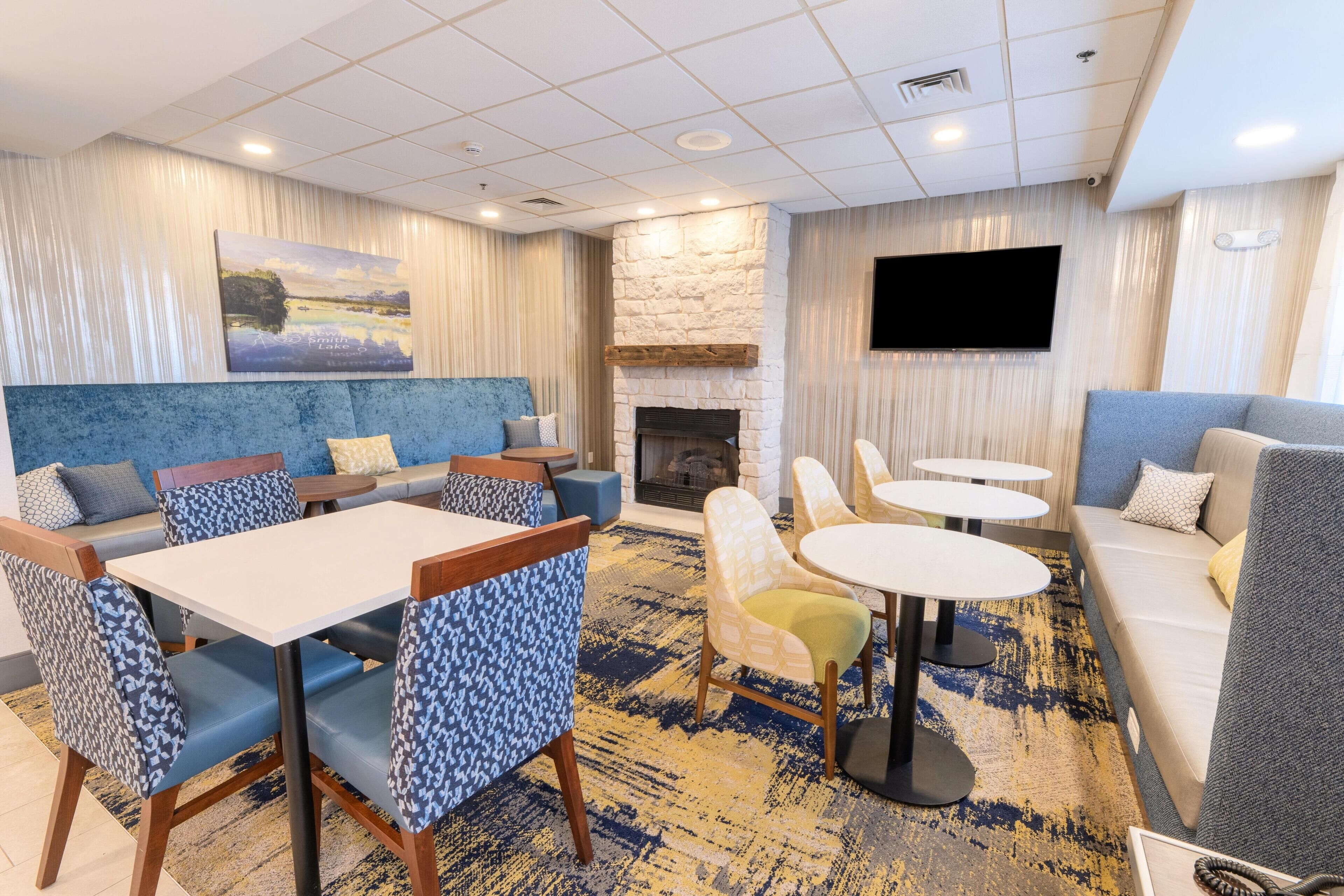 A modern hotel lobby with a stone fireplace, a large TV mounted on the wall, and a seating area with sofas and tables.