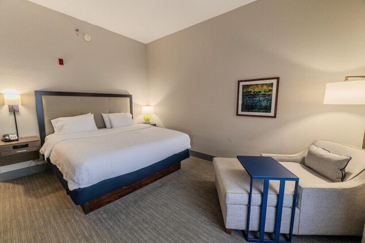 A hotel room with a bed, bedside table, lamp, and a small table with a chair.