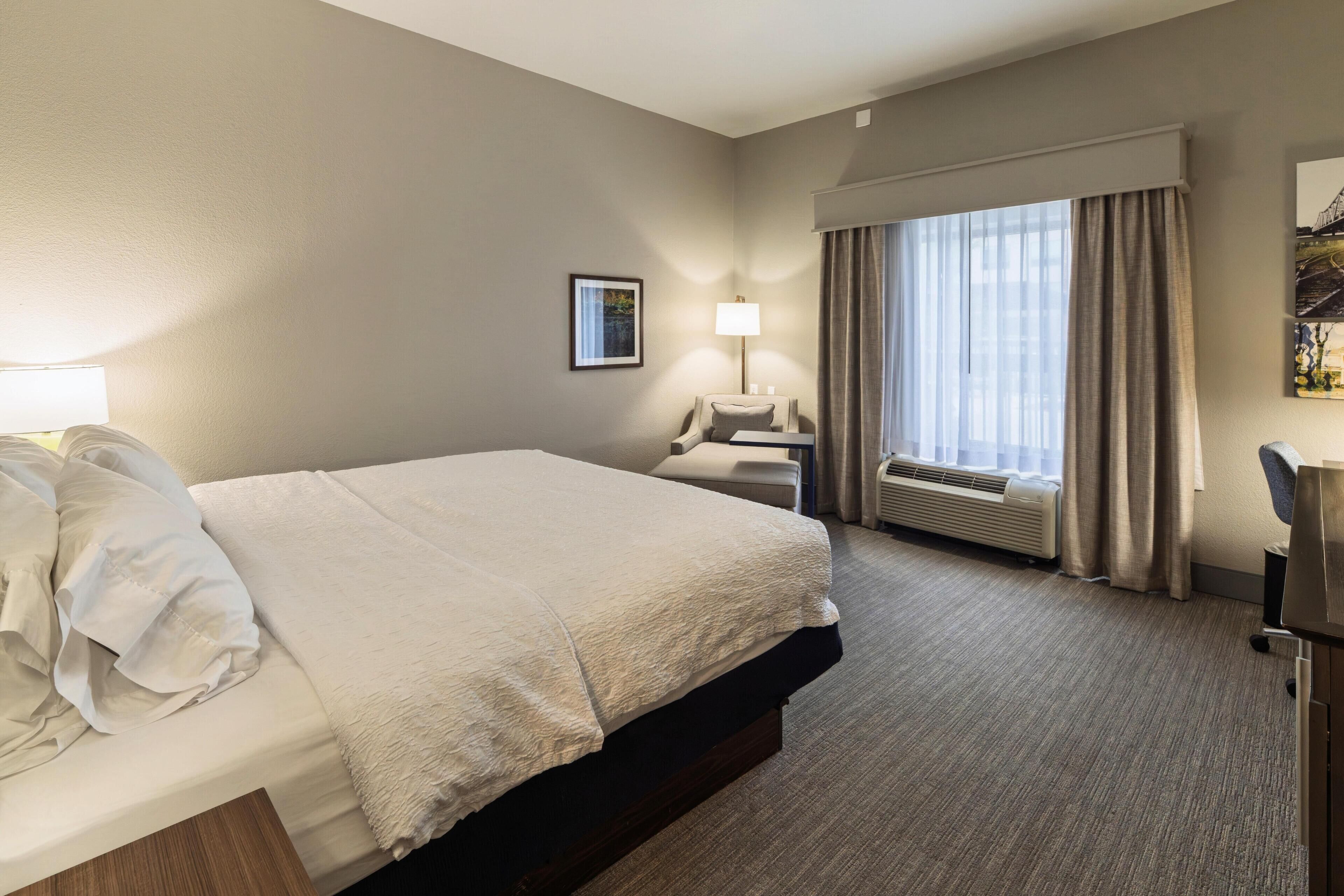 Habitación de hotel con una cama grande, un escritorio, una silla, una lámpara y una ventana con cortinas.