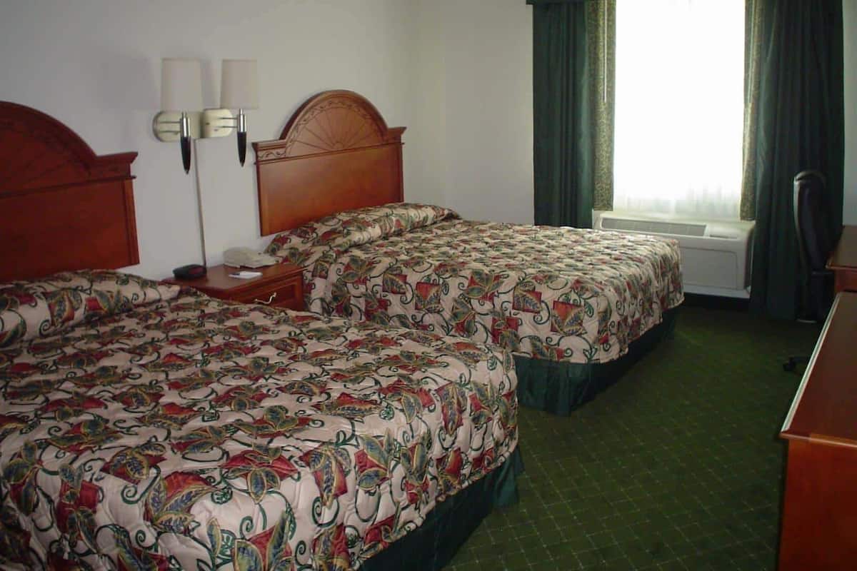 A hotel room with two beds, a nightstand, a lamp, a window with curtains, and a radiator.