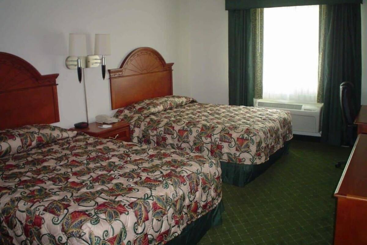 A hotel room with two beds, a nightstand, a lamp, a window with curtains, and a radiator.