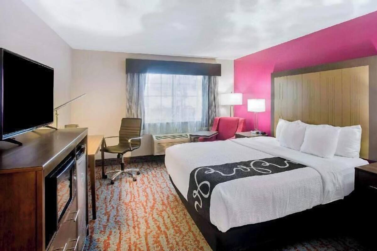 A hotel room with a bed, a desk with a TV, a chair, a lamp, and a window with curtains.