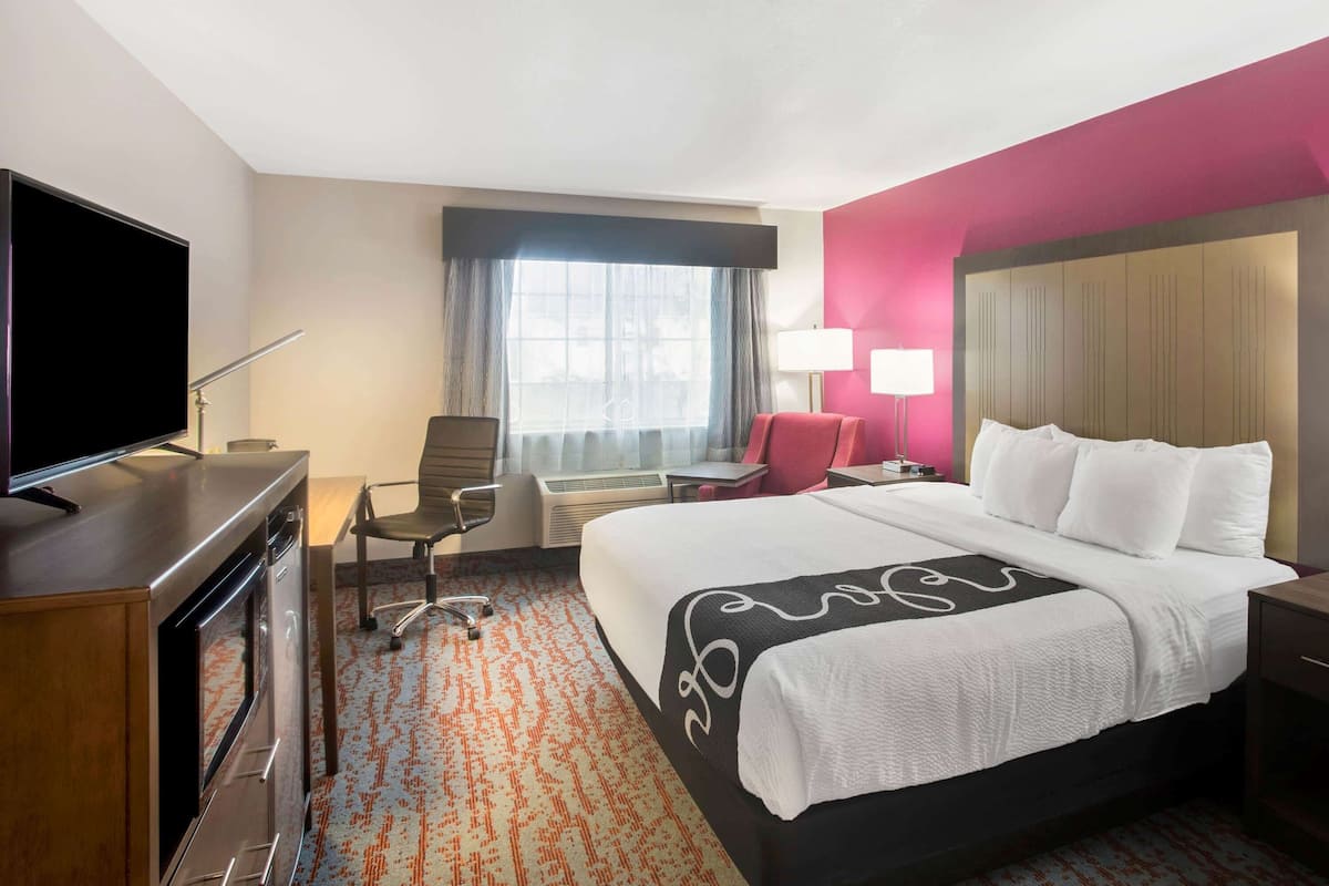 A hotel room with a large bed, a desk with a flat-screen TV, a chair, a desk lamp, and a window with curtains.