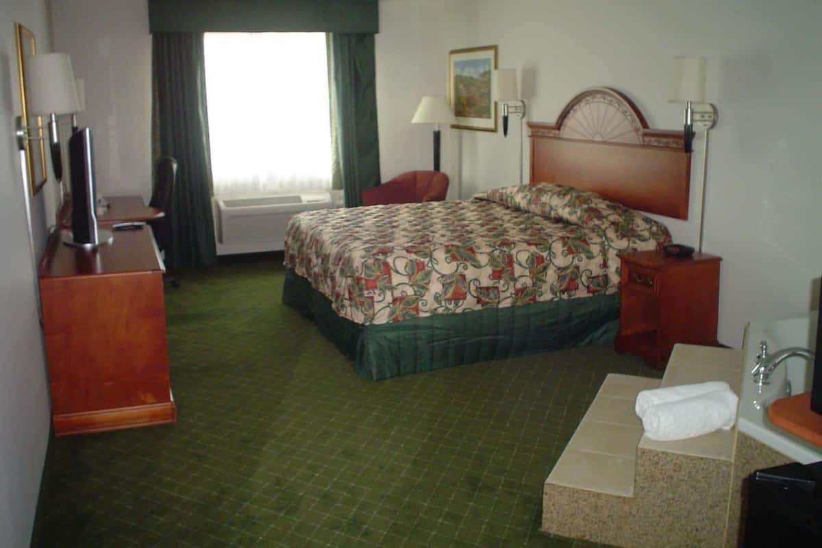 A hotel room with a bed, a desk, a chair, a desk lamp, a bedside table, a mirror, a window with curtains, and a bathroom with a sink and a towel.