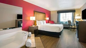 Habitación de hotel con una cama grande, un escritorio independiente, una silla de escritorio, una lámpara de escritorio, un televisor de pantalla plana, un ventanal con cortinas y un baño con bañera, además de dos l ámparas de noche.