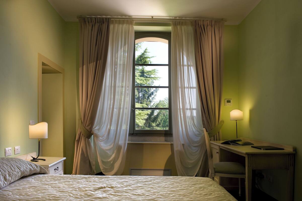 Un dormitorio con cama, escritorio, silla, ventana con cortinas y lámpara.