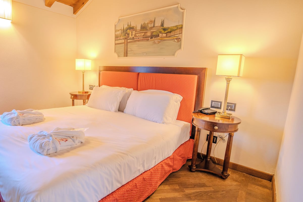 Una cama bien hecha con sábanas blancas y dos almohadas, un cabecero de madera, dos mesitas de noche con lámparas y un cuadro en la pared.