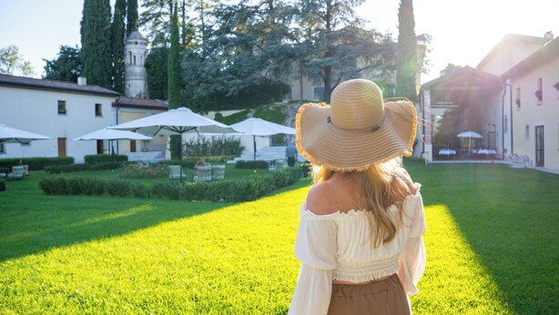 Una persona con blusa blanca y falda marrón parada en un jardín bien cuidado, con sombrillas blancas y un edificio al fondo.