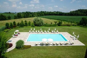 Una piscina rectangular con solárium de piedra, rodeada de sillas de descanso y sombrillas, ubicada en un paisaje verde exuberante con campos y árboles al fondo.