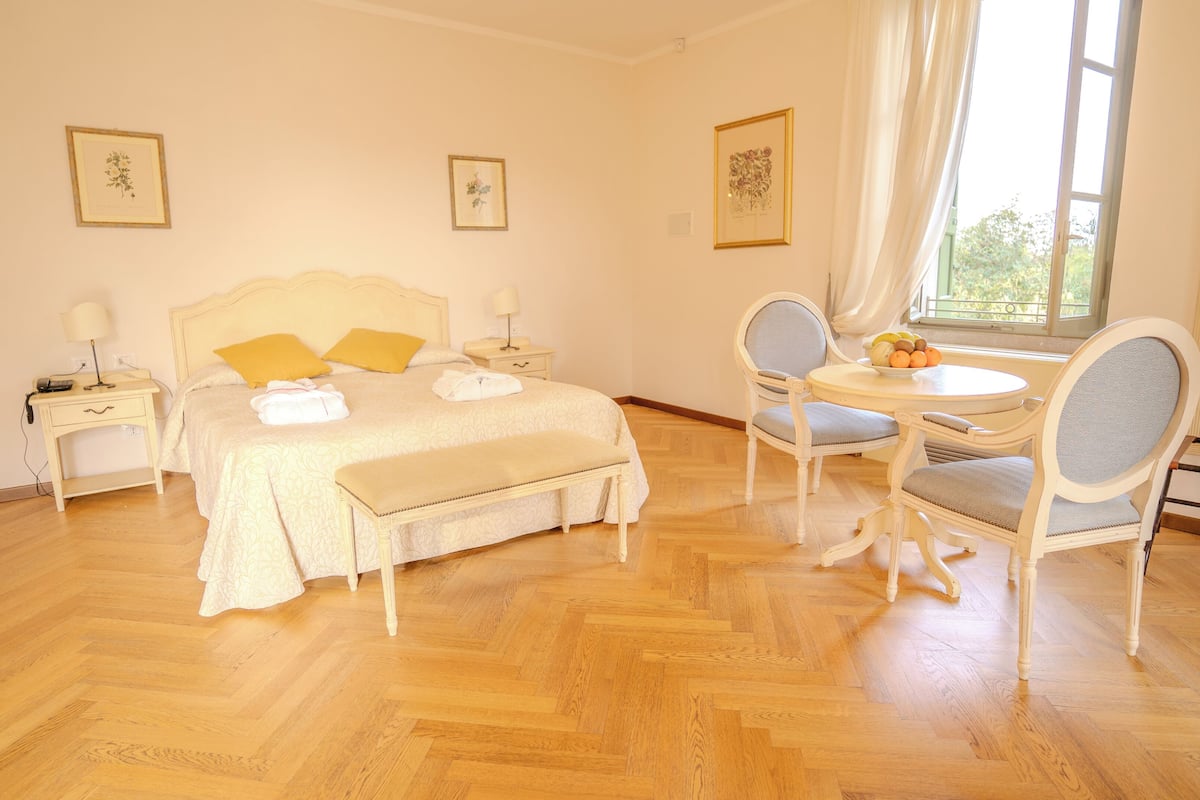 Un dormitorio con cama, mesitas de noche, un banco, una mesa redonda con sillas y cuadros en las paredes.