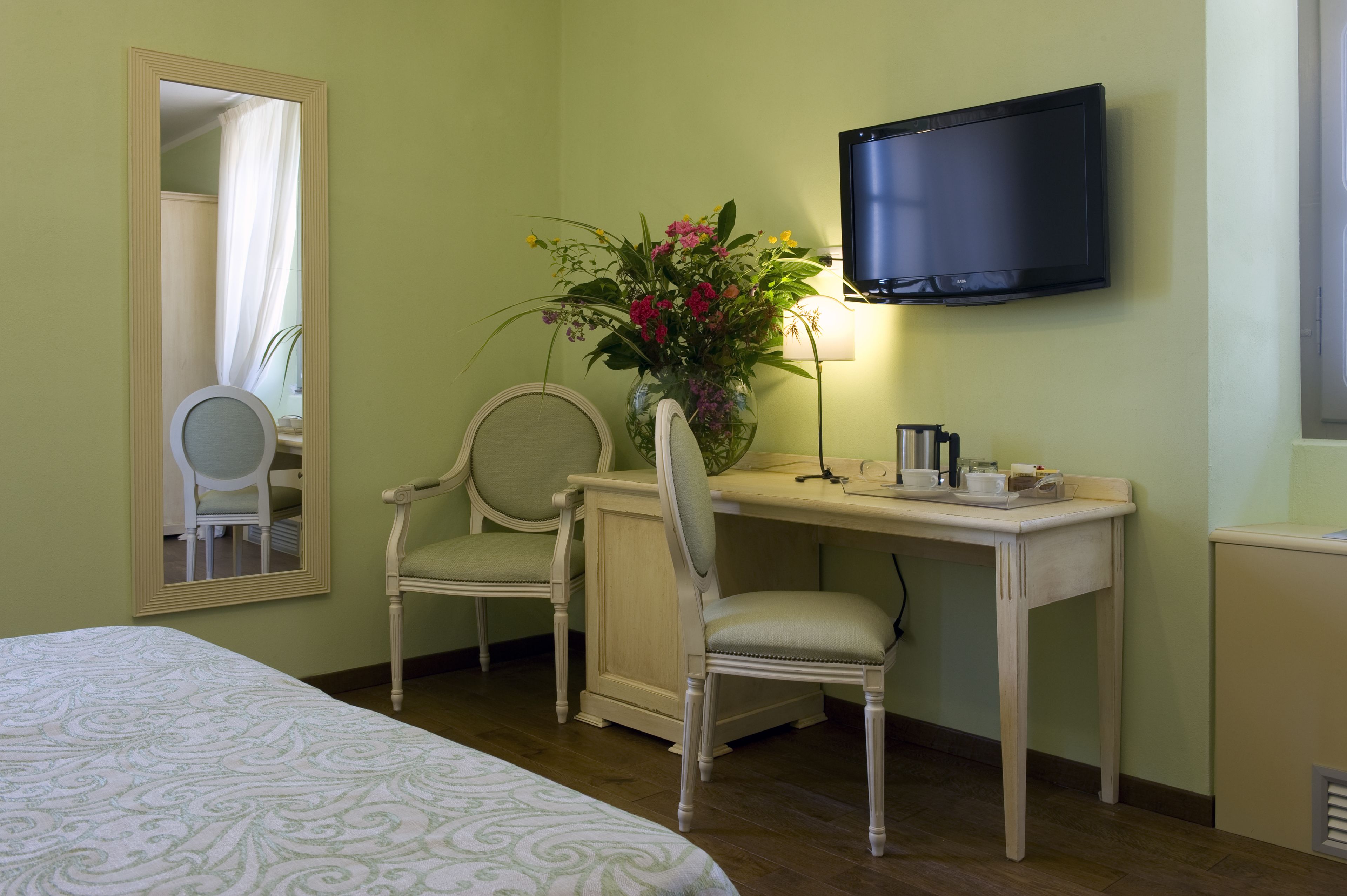 A hotel room with a bed, two chairs, a desk with a TV, a vase with flowers, and a mirror.
