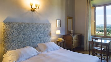 Una cama bien tendida con sábanas blancas, una lámpara de noche, un aparador de madera y una silla junto a una ventana con vistas.