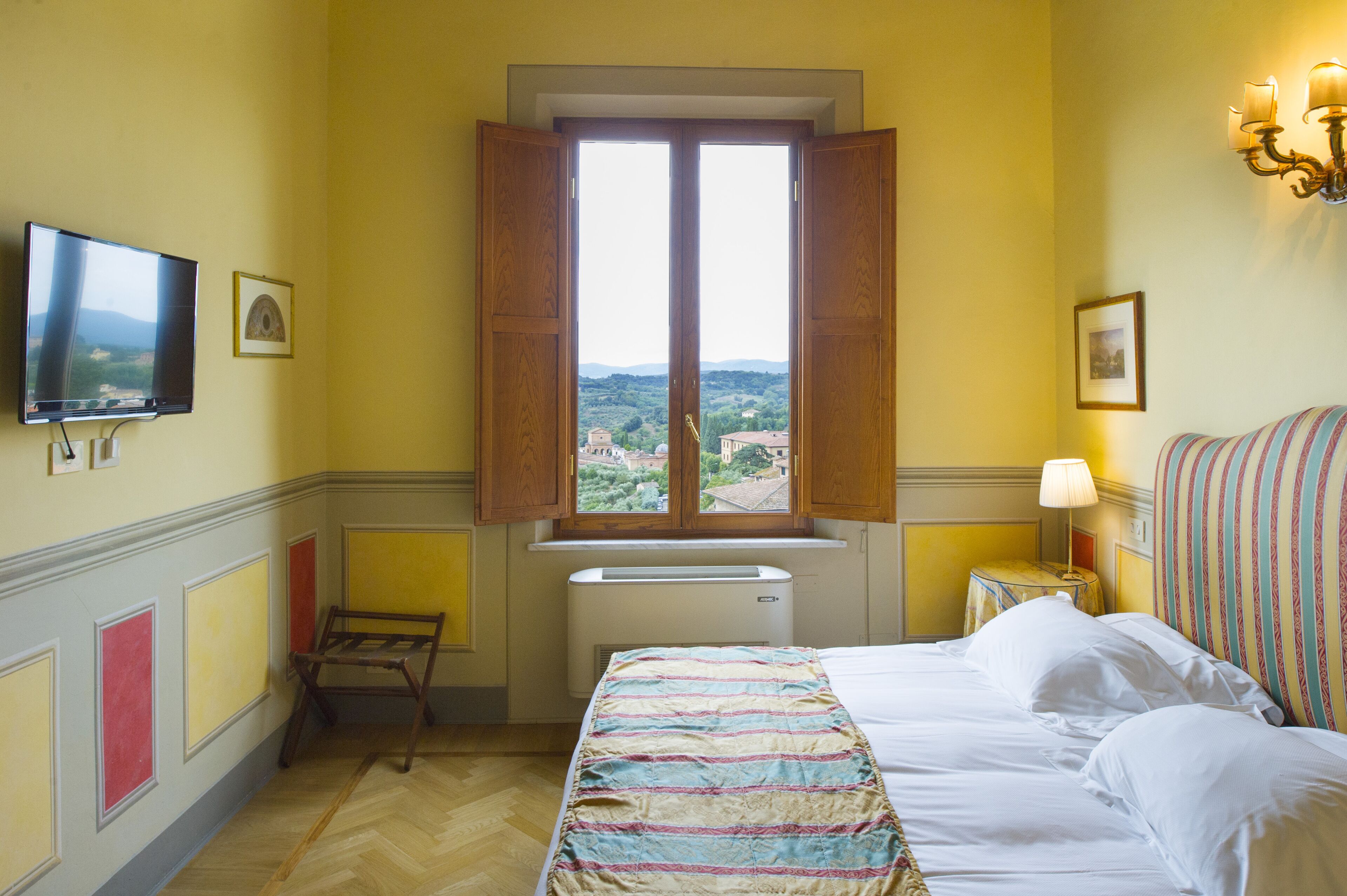 A hotel room with a bed, a window with open shutters, a television, and a wall-mounted lamp.