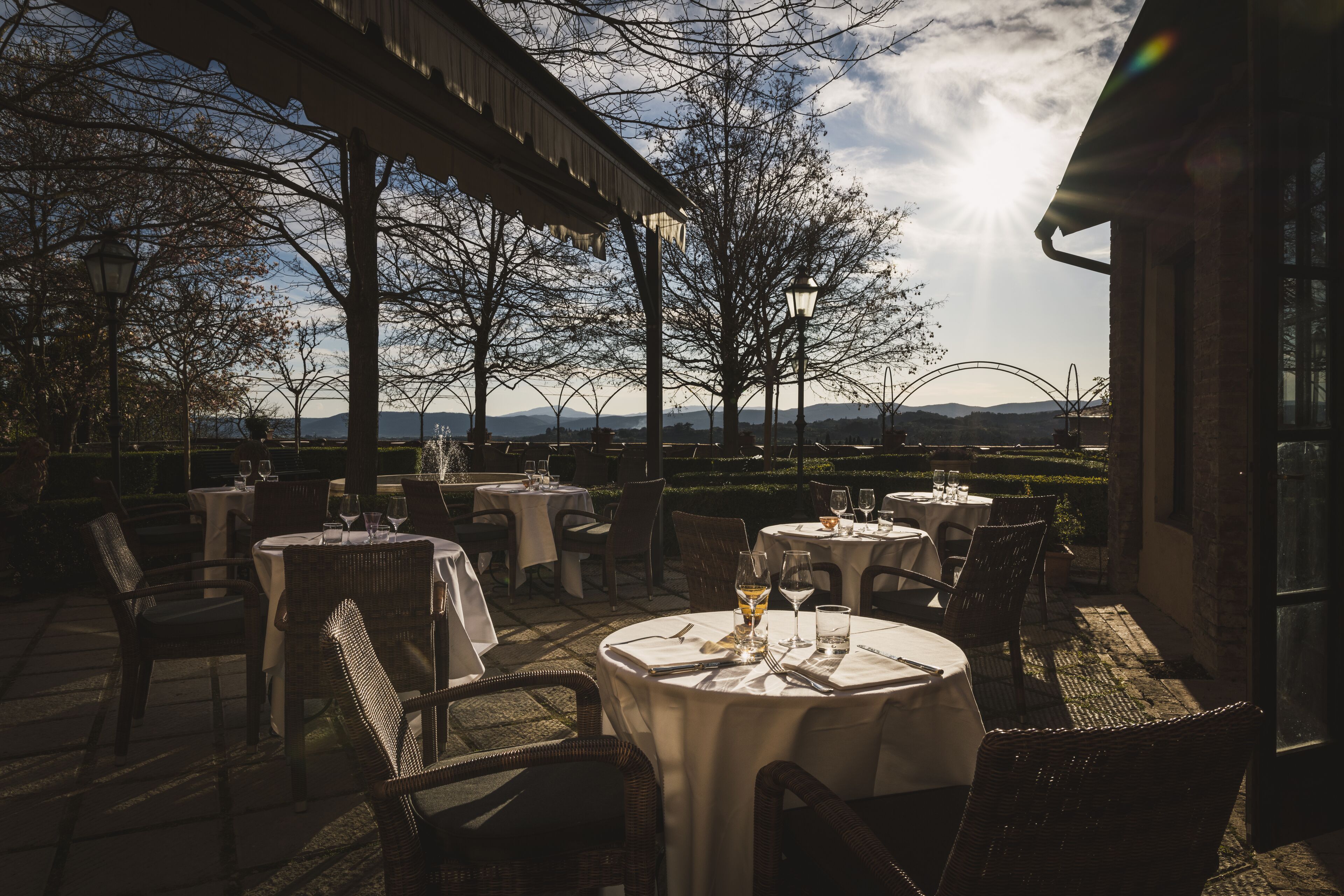 An outdoor dining area with round tables and wicker chairs, set against a backdrop of trees and distant mountains.