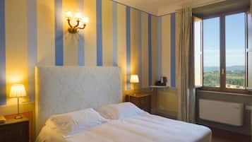 Una camera d'albergo con un letto grande, un comodino, una lampada, una cassettiera, una finestra con tende e vista sulla campagna.