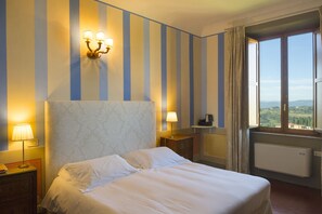 A hotel room with a large bed, a nightstand, a lamp, a dresser, a window with curtains, and a view of the countryside.
