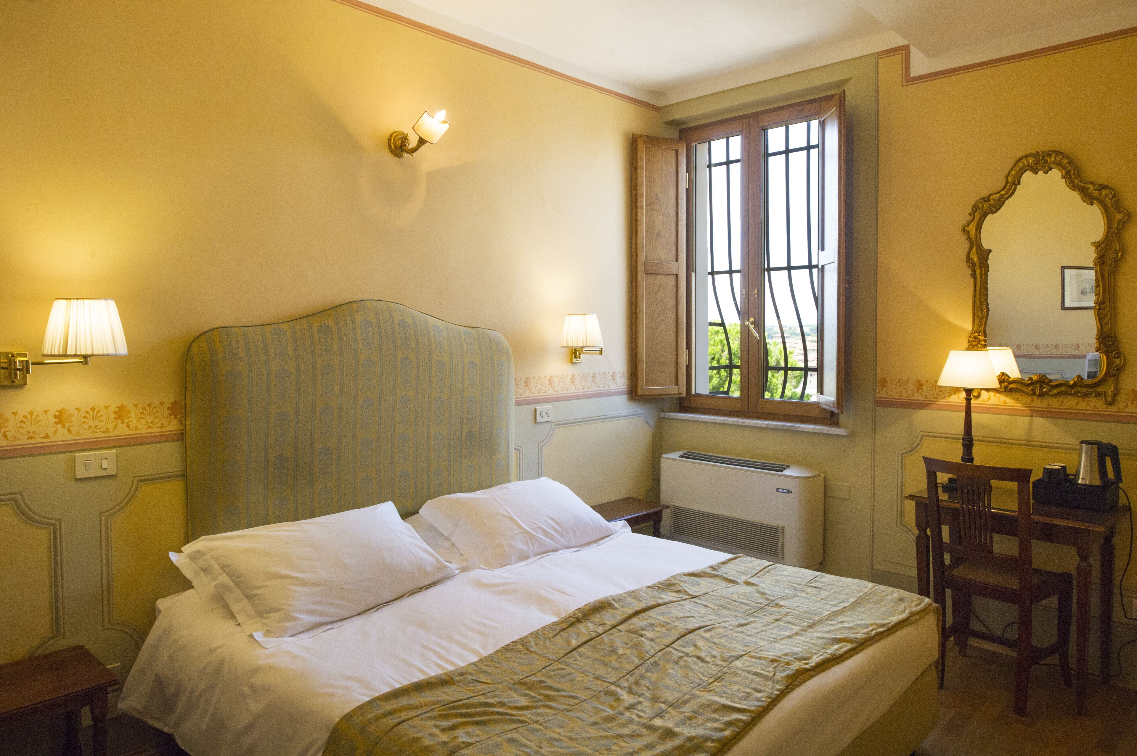 Una camera d'albergo con un letto, due lampade ai lati, una sedia in legno, una scrivania con lampada, un termosifone e una finestra con inferriate.