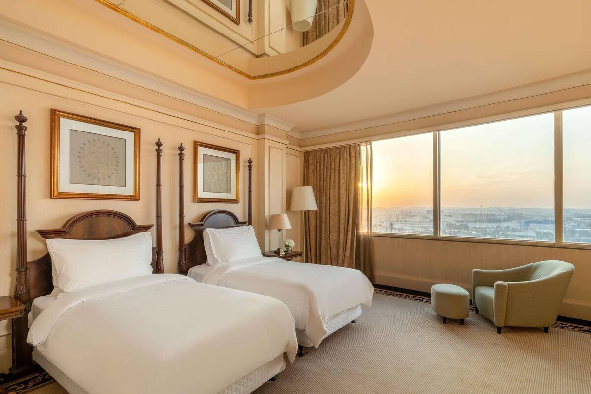 A hotel room with two beds, a large window with a city view, a chair, and a lamp.