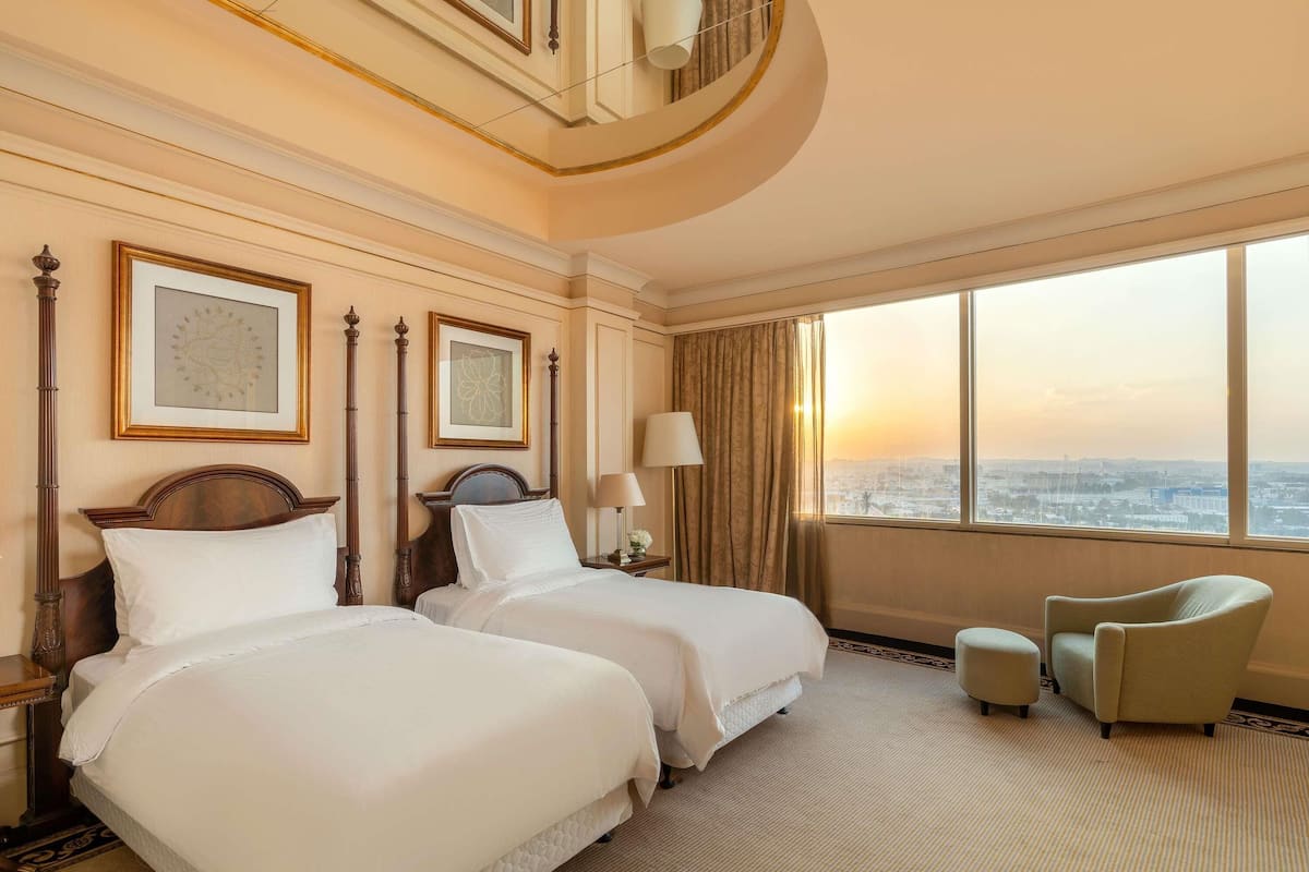 Habitación de hotel con dos camas, un ventanal amplio con vista a la ciudad, una silla y una lámpara.