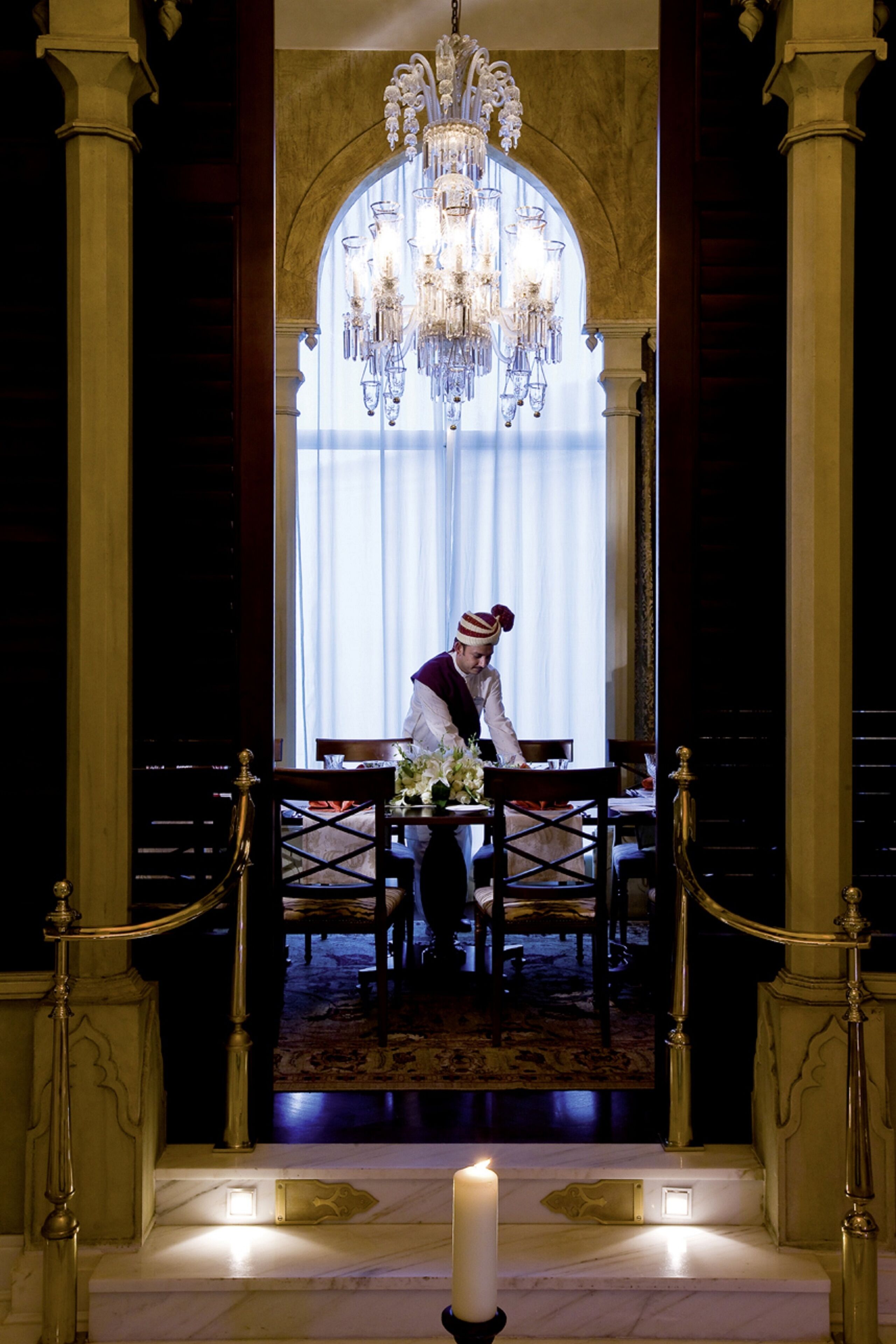 A grand hall with a chandelier, a white candle, and a table set for a meal.