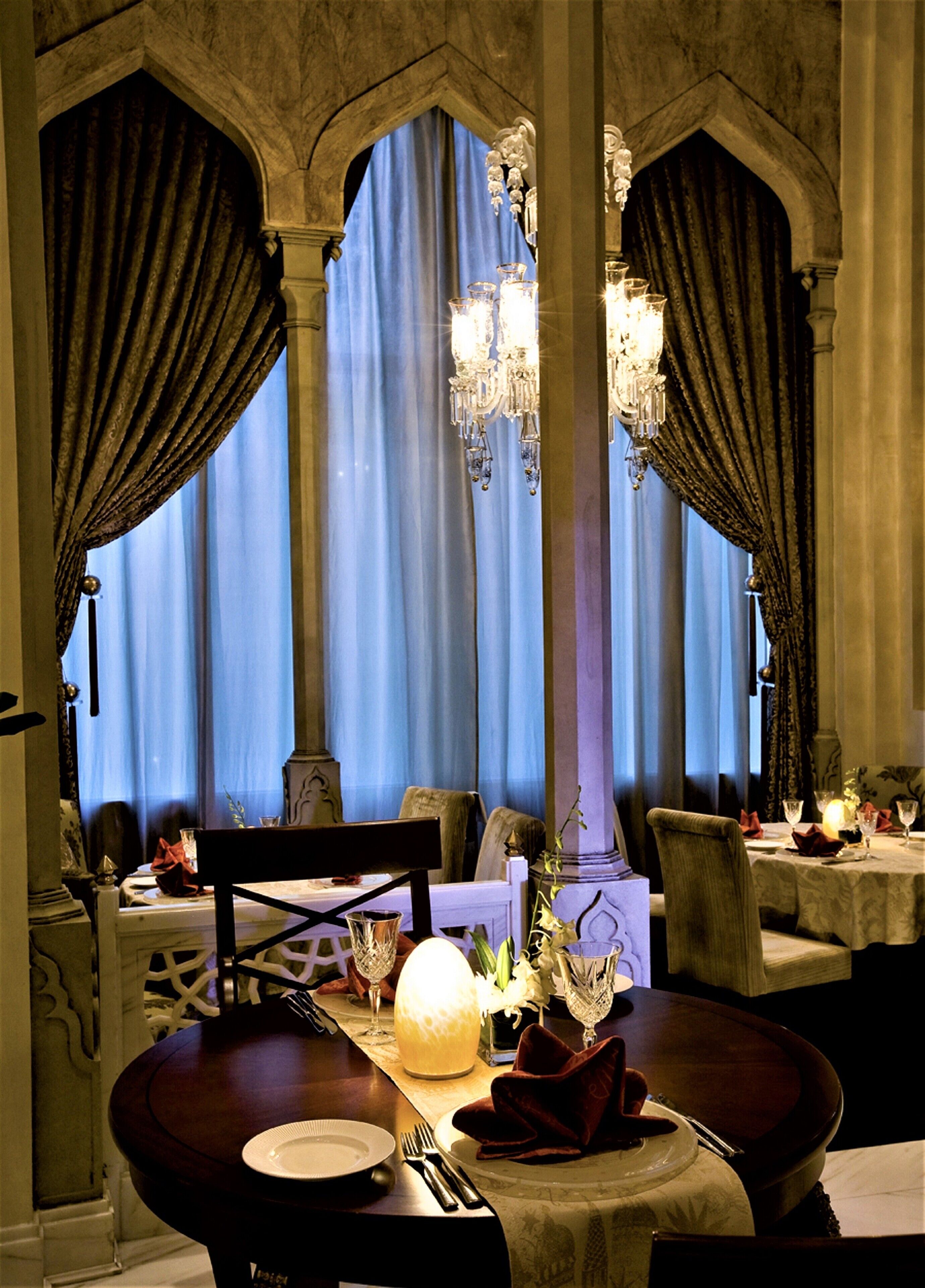 A dining area with a table set for two, ornate chandeliers, and draped curtains.