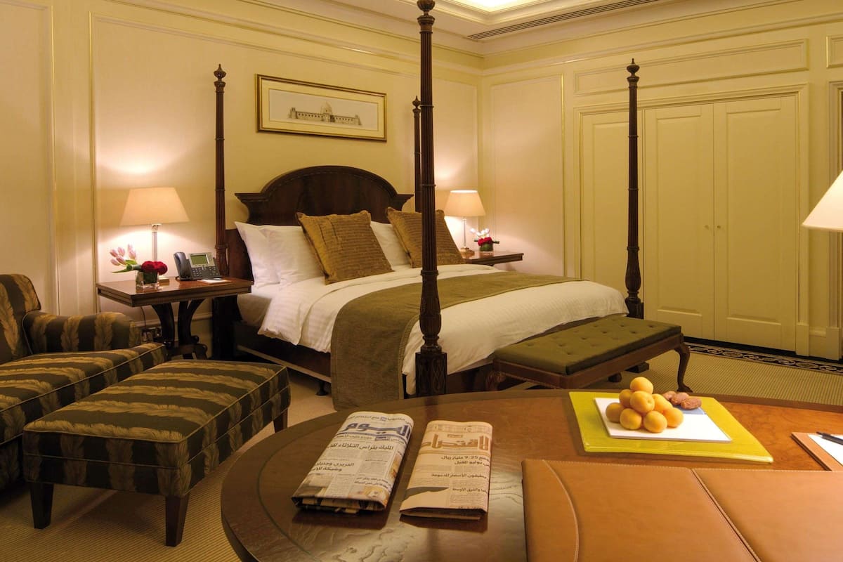 A four-poster bed with a headboard, a bedside table with a lamp, a chair, a small table with a newspaper, and a fruit basket.