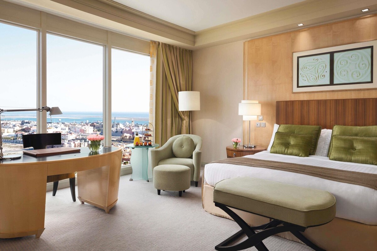 Una habitación de hotel con escritorio, un sillón cómodo, una cama con almohadas verdes, una mesita de noche con lámpara y vistas a la ciudad.