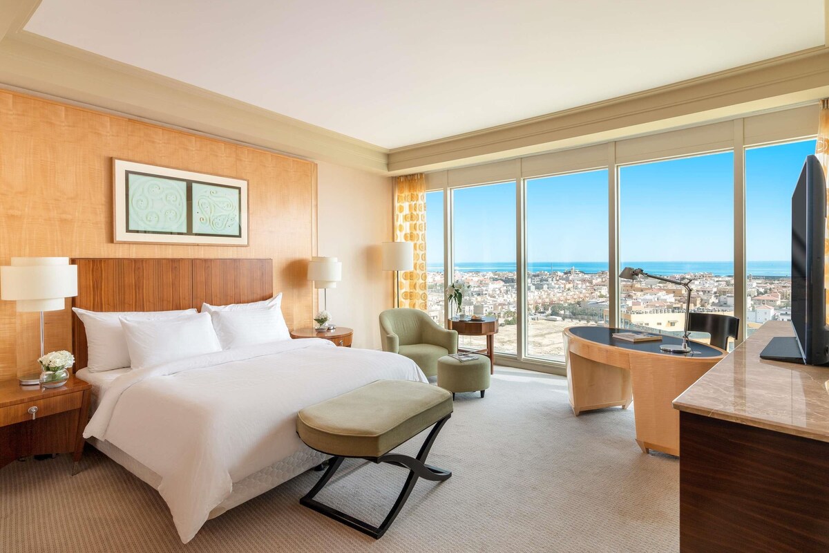 A hotel room with a large bed, a desk, a chair, and a view of the city and sea.