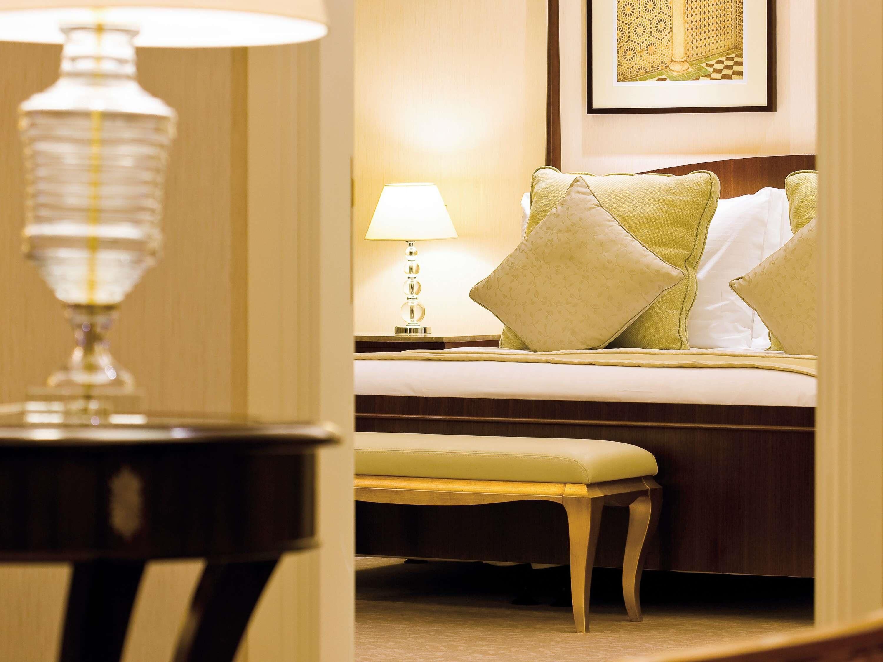 A hotel room with a bed, a bedside table with a lamp, a bench, and a framed picture on the wall.