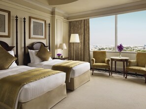 A hotel room with two beds, a sitting area with a chair, a small table, and a view of the city. - Mövenpick Hotel Al Khobar (Al Khobar)