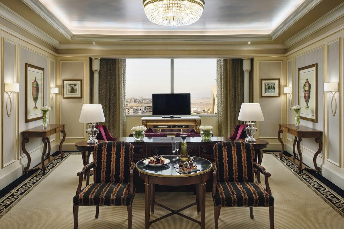 A hotel room with a large window, a chandelier, a dining table with chairs, and a flat-screen TV.