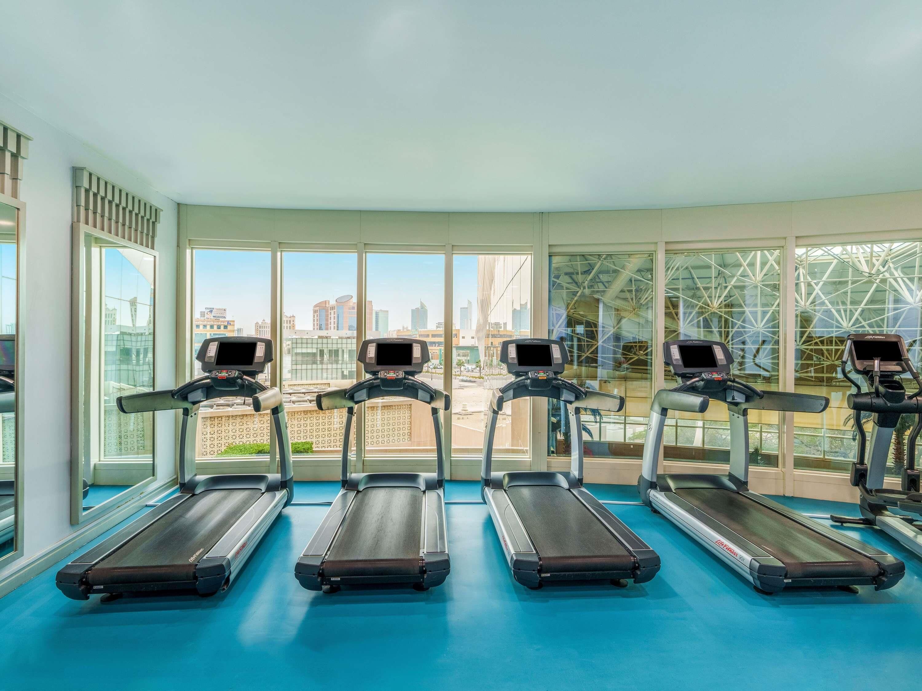 Une salle de sport avec quatre tapis de course alignés en rangée, de grandes fenêtres donnant sur la ville et un sol bleu.