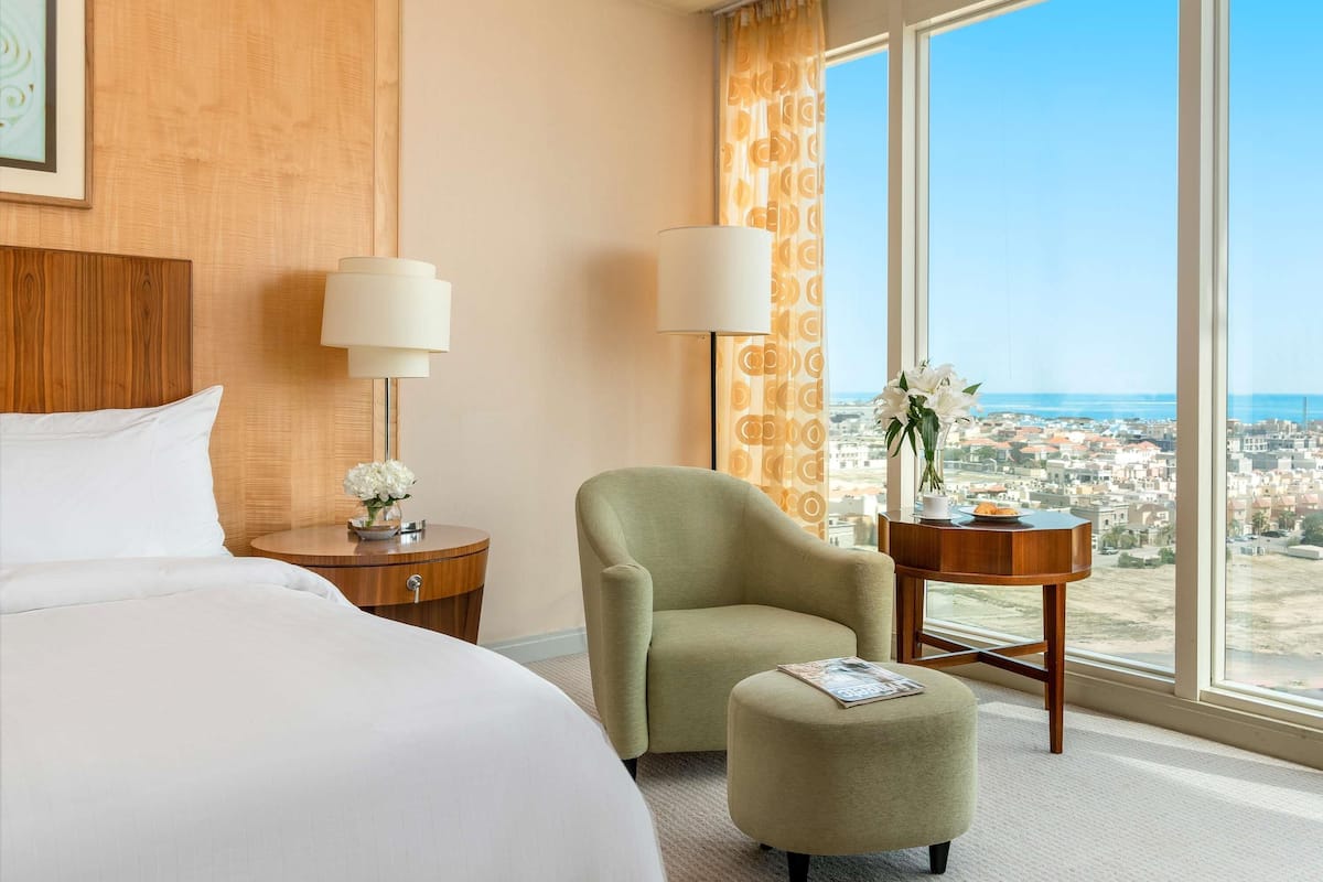 A hotel room with a bed, two nightstands, a green armchair, a round ottoman, and a large window with a city view.
