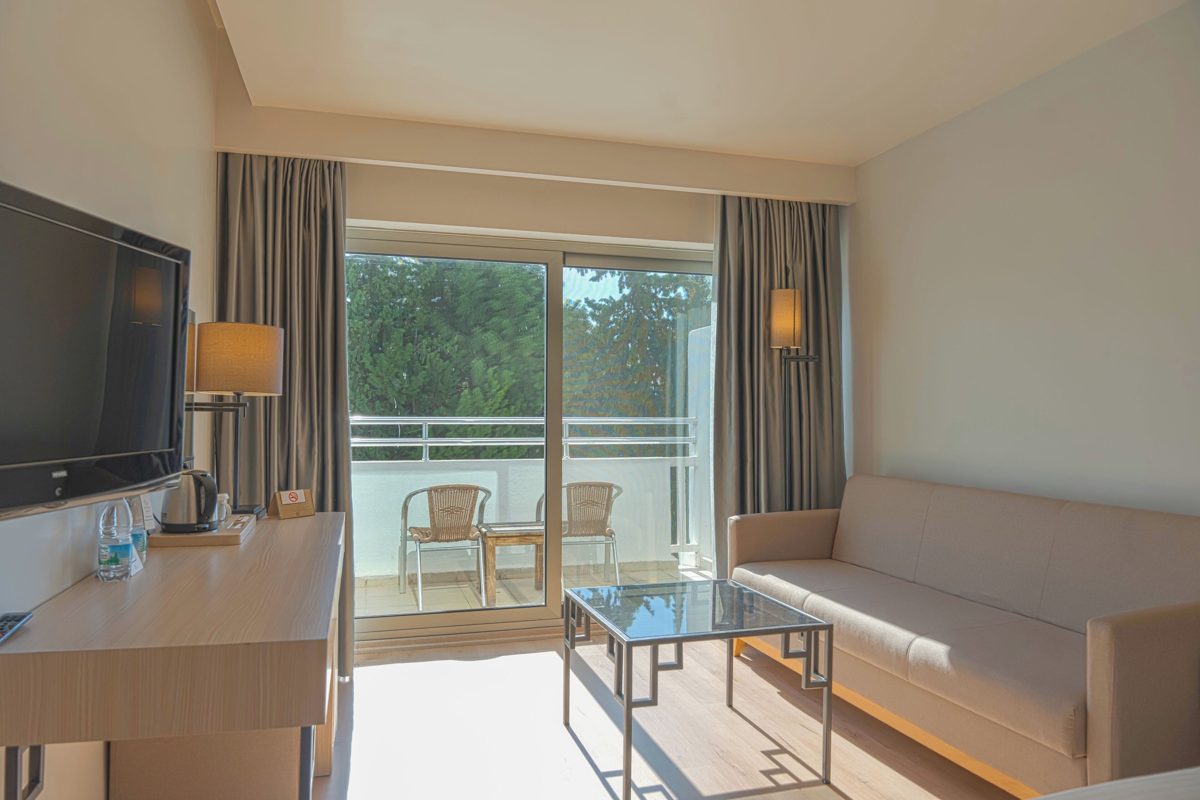 A modern hotel room with a sofa, a glass coffee table, a flat-screen TV, a desk with a lamp, and a balcony with chairs.