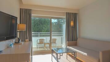 A modern hotel room with a sofa, a glass coffee table, a flat-screen TV, a desk with a lamp, and a balcony with chairs.