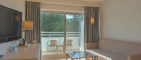 A modern hotel room with a sofa, a glass coffee table, a flat-screen TV, a desk with a lamp, and a balcony with chairs.