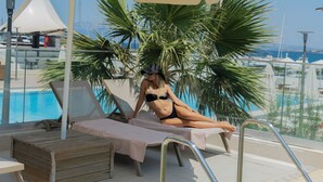 Zona de piscina con una tumbona, una palmera y una sombrilla blanca.