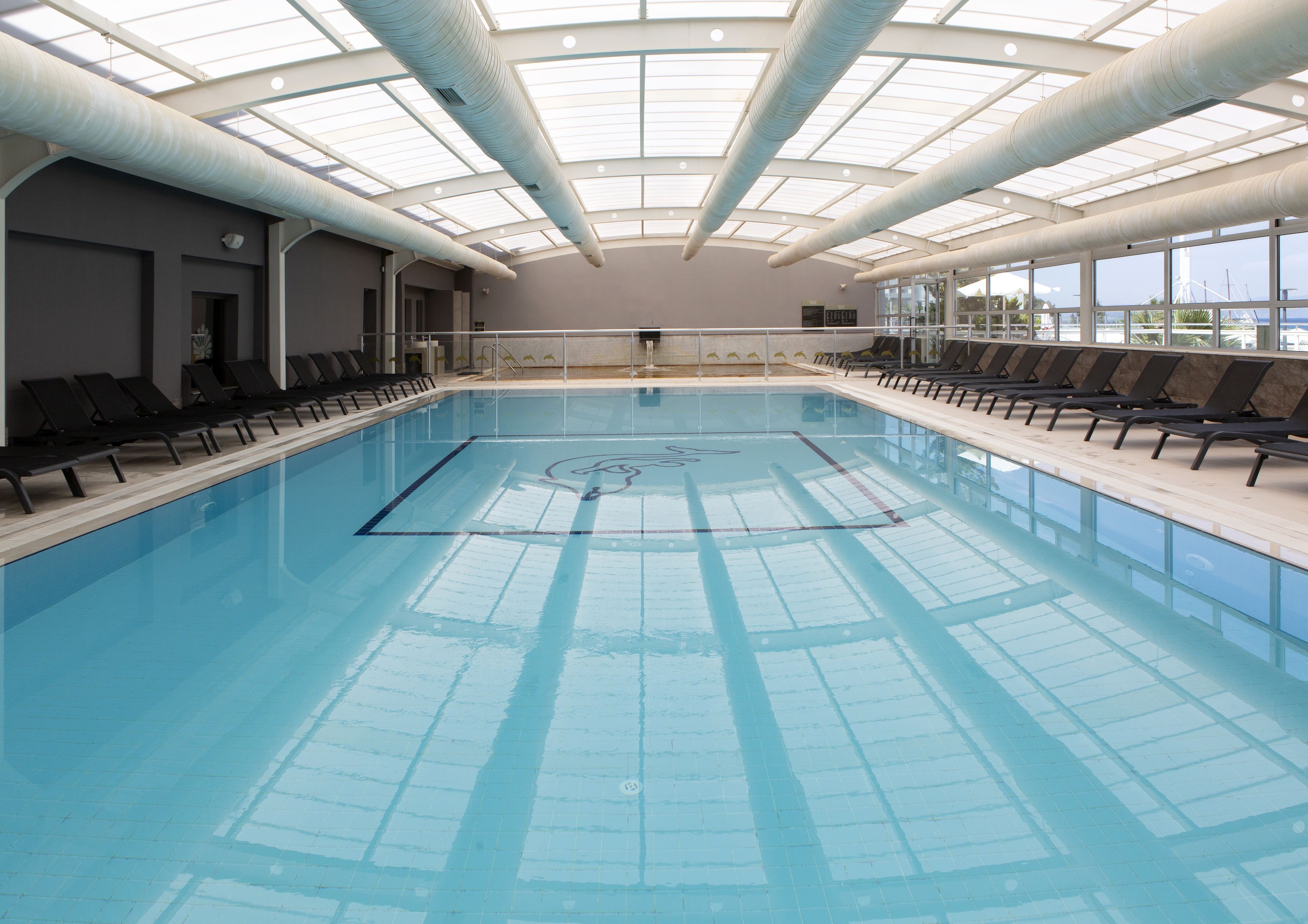 An indoor swimming pool with clear lanes, sunlit by skylights, and equipped with sun loungers along the sides.