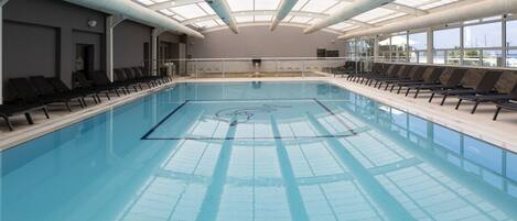 An indoor swimming pool with clear lanes, sunlit by skylights, and equipped with sun loungers along the sides.