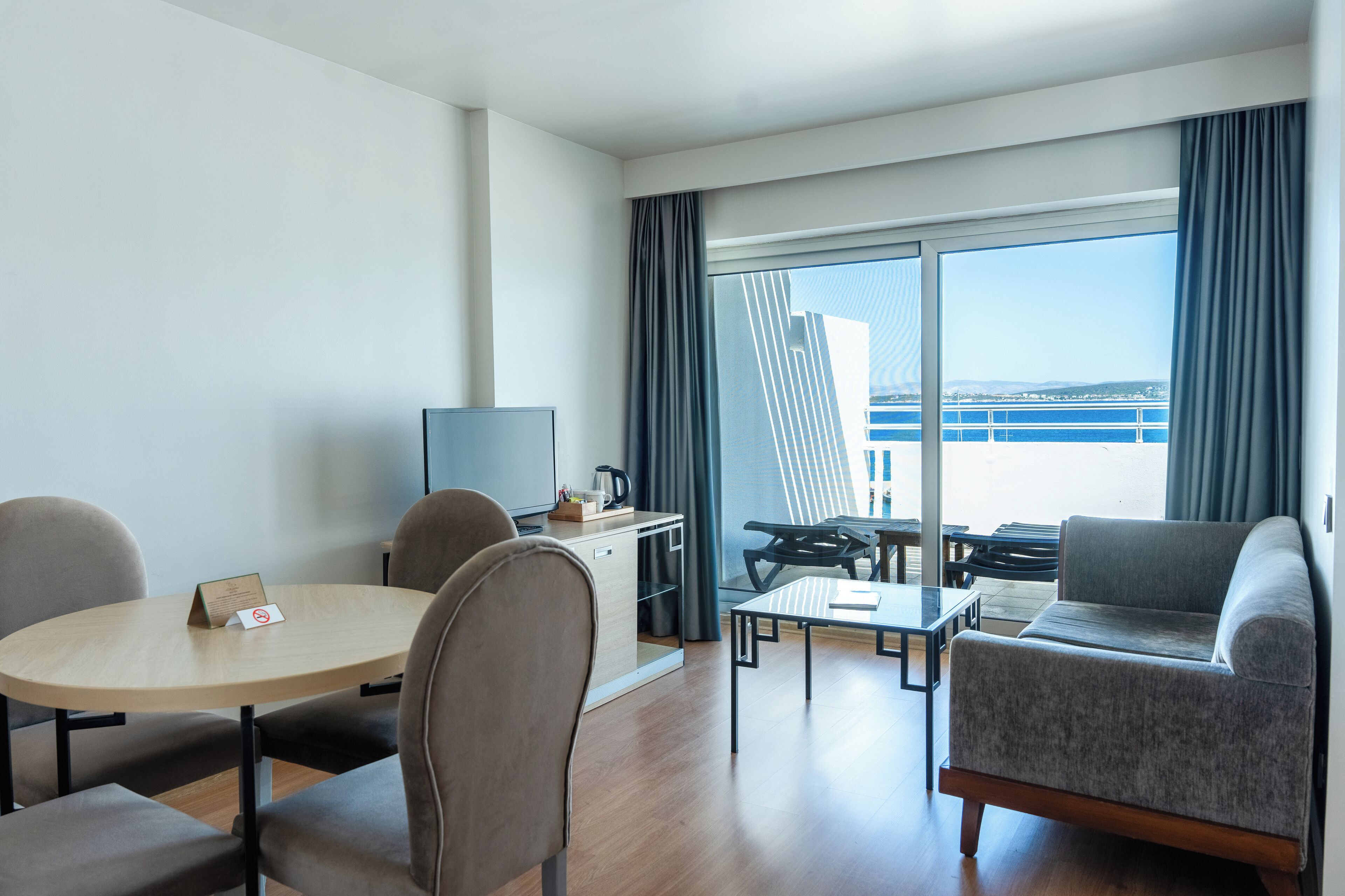 A modern hotel room with a round table, two chairs, a sofa, a TV, and a balcony with a view of the sea.