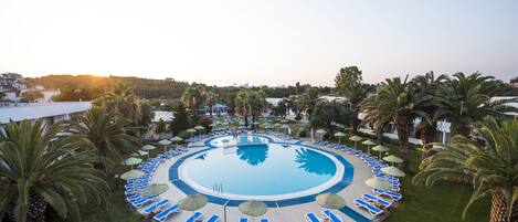 A circular pool with lounge chairs and umbrellas, surrounded by palm trees and a hilly landscape.