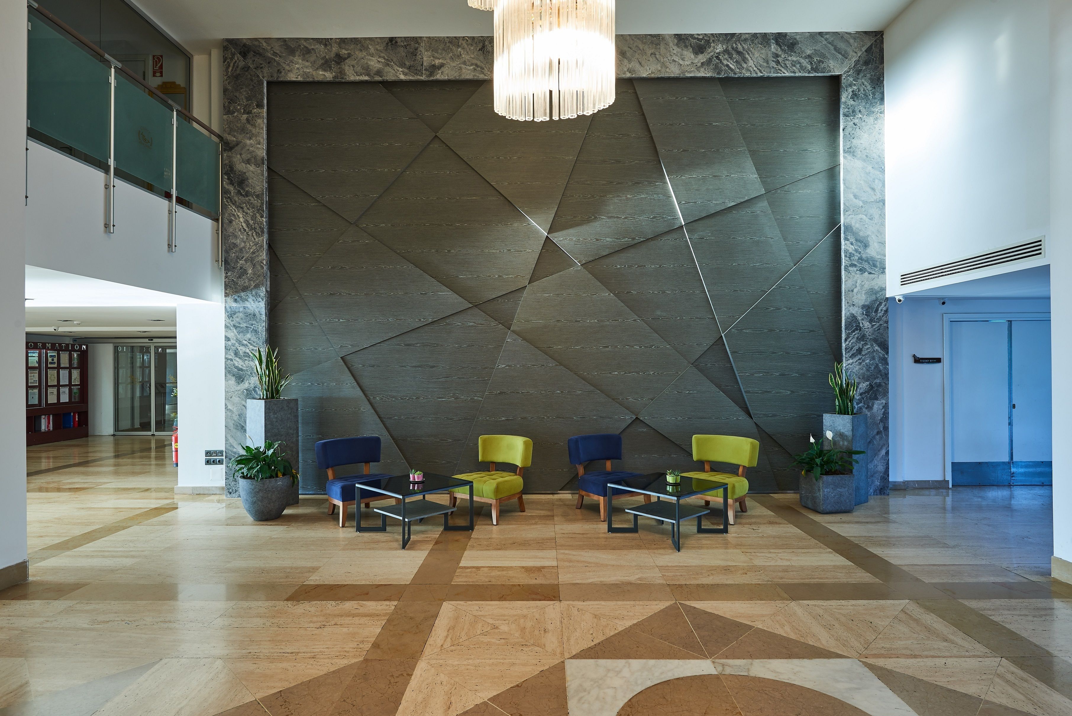 A lobby with a geometric patterned wall, a chandelier, and seating with potted plants.