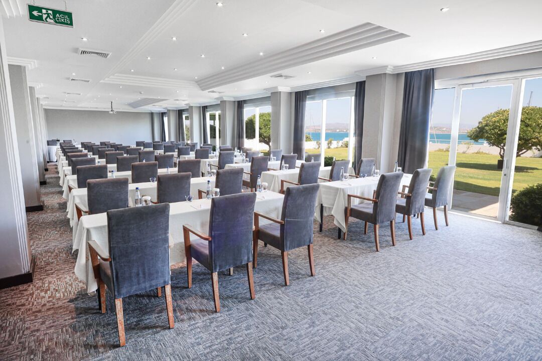 Een vergaderruimte met rijen tafels en stoelen, grote ramen met uitzicht op de zee, en een vloerbedekking.