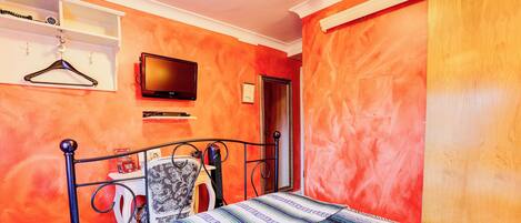 A bedroom with a bed, a TV, a wardrobe, and a shelf with a hanging garment.