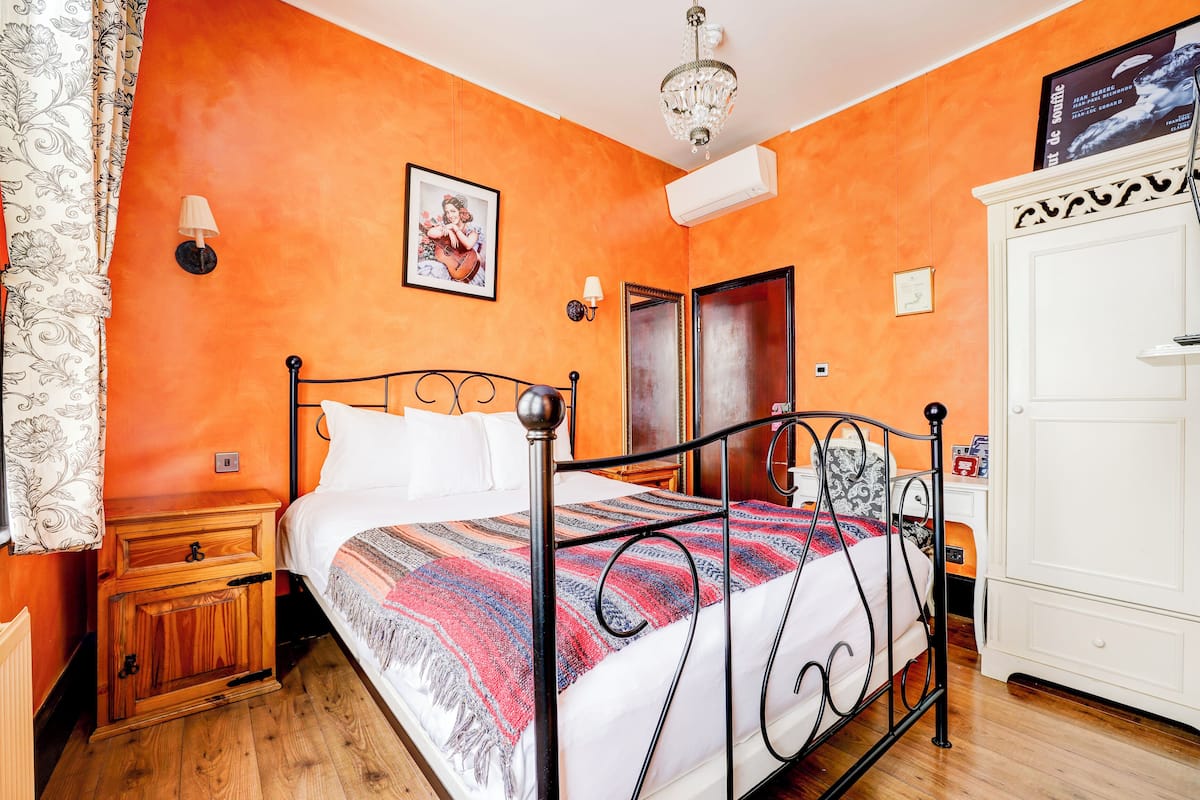 A bedroom with an orange wall, a bed with a patterned blanket, a wooden nightstand, a white wardrobe, and a chandelier.