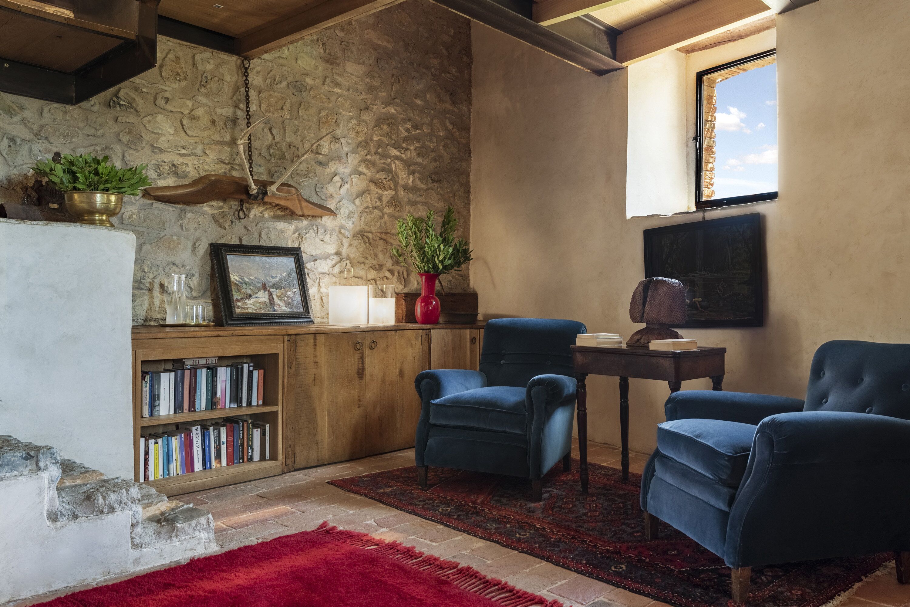 A cozy reading nook with a stone wall, a bookshelf, a red rug, and two blue armchairs.