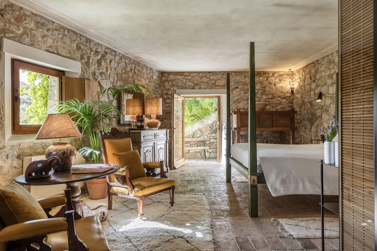 Una habitación rústica con paredes de piedra, una cama, una silla, una mesa con una lámpara y una planta en maceta.