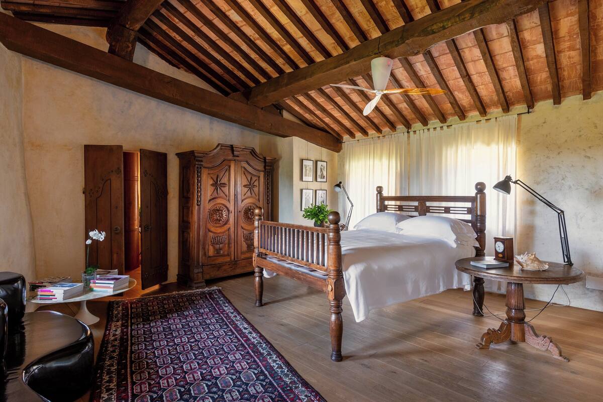Un dormitorio con techo de madera, una cama, un banco de madera, un escritorio con libros y una alfombra.