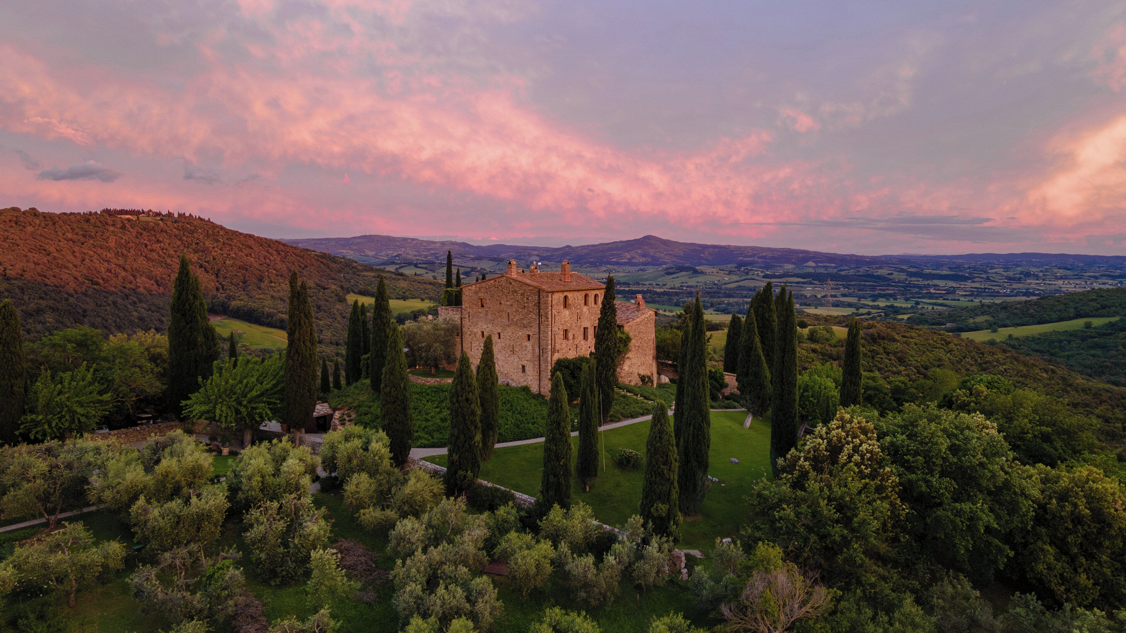 Un castillo rodeado por un jardín cuidado con altos cipreses, con colinas onduladas y un cielo al atardecer de fondo.