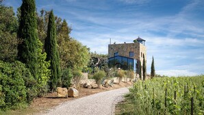 A stone tower with a glass extension, surrounded by greenery and a gravel path. - Castello Di Vicarello (Cinigiano)